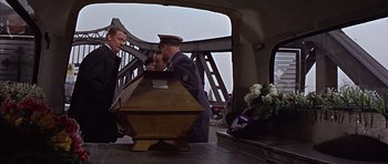 Movie still from “Funeral in Berlin” (1966), directed by Guy Hamilton – A man in a military uniform standing next to a wooden casket; Medium shot, High angle