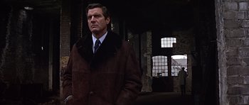 Movie still from “Funeral in Berlin” (1966), directed by Guy Hamilton – A man in a brown coat and tie standing in front of a brick wall; Medium shot, Low angle