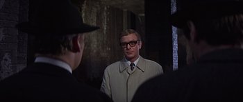 Movie still from “Funeral in Berlin” (1966), directed by Guy Hamilton – A man wearing a suit and tie standing next to a man wearing a hat; Close Up shot, Over the shoulder angle