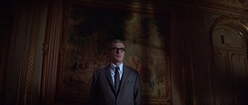 Movie still from “Funeral in Berlin” (1966), directed by Guy Hamilton – A man in a suit and tie standing in front of a painting; Medium shot, Low angle