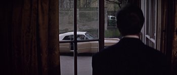Movie still from “Funeral in Berlin” (1966), directed by Guy Hamilton – A man standing in front of a car in a parking lot; Wide shot, Over the shoulder angle
