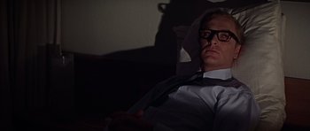 Movie still from “Funeral in Berlin” (1966), directed by Guy Hamilton – A man in a white shirt and tie sitting in a chair; Medium shot, Low angle