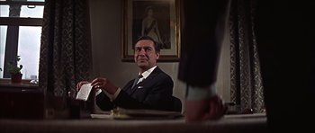 Movie still from “Funeral in Berlin” (1966), directed by Guy Hamilton – A man sitting at a table in front of a painting; Medium shot, Over the shoulder angle