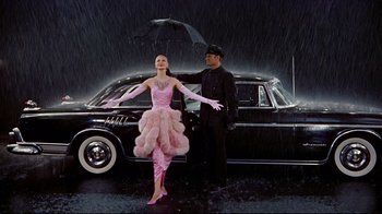 Movie still from “Funny Face” (1957), directed by Stanley Donen – A man and a woman standing in front of an old car in the rain; Wide shot, Low angle
