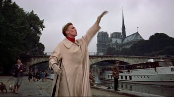 Movie still from “Funny Face” (1957), directed by Stanley Donen – An older woman in a trench coat waves at the crowd; Wide shot, Low angle