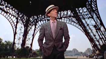 Movie still from “Funny Face” (1957), directed by Stanley Donen – A man in a suit and hat standing under an iron structure; Medium shot, Low angle