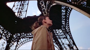 Movie still from “Funny Face” (1957), directed by Stanley Donen – A woman in a tan jacket standing in front of the eiffel tower; Medium shot, Low angle