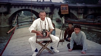 Movie still from “Funny Face” (1957), directed by Stanley Donen – A man sitting in a chair holding a camera in front of two other people; Medium shot, High angle