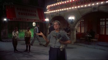 Movie still from “Funny Face” (1957), directed by Stanley Donen – An older woman holding a dog in front of a building; Wide shot, Low angle