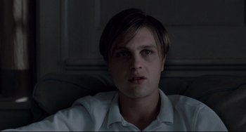 Movie still from “Funny Games” (2007), directed by Michael Haneke – A young man with long brown hair sitting on a couch; Close Up shot, High angle