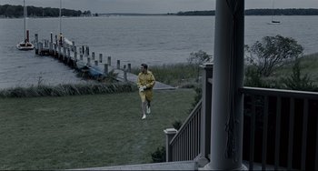 Movie still from “Funny Games” (2007), directed by Michael Haneke – A man in a raincoat walking on the grass by the water; Extreme Wide shot, High angle