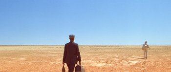 Movie still from “Gallipoli” (1981), directed by Peter Weir – A man in a suit and hat holding a suitcase and walking across a barren field; Extreme Wide shot, Over the shoulder angle