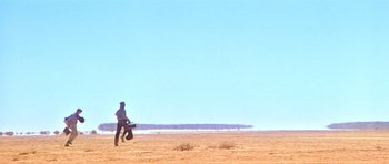 Movie still from “Gallipoli” (1981), directed by Peter Weir – A man standing on top of a sandy beach holding a surfboard; Extreme Wide shot, Low angle