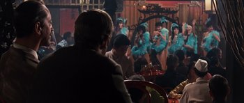 Movie still from “Gambit” (1966), directed by Ronald Neame – A group of people sitting at a table in front of a crowd; Wide shot, Over the shoulder angle