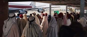 Movie still from “Gambit” (1966), directed by Ronald Neame – A group of people standing in front of an airplane on a runway; Wide shot, Low angle