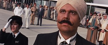 Movie still from “Gambit” (1966), directed by Ronald Neame – A man in a suit and a turban is standing in front of a group of people; Close Up shot, Low angle