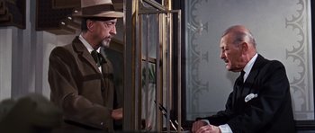 Movie still from “Gambit” (1966), directed by Ronald Neame – A man in a suit and hat talking to another man in a suit and hat; Medium shot, Over the shoulder angle