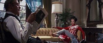 Movie still from “Gambit” (1966), directed by Ronald Neame – A woman sitting on a couch in a living room; Wide shot, Over the shoulder angle
