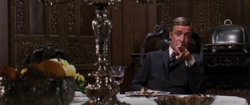 Movie still from “Gambit” (1966), directed by Ronald Neame – A man sitting at a dinner table with a glass of wine; Medium shot, Over the shoulder angle