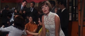 Movie still from “Gambit” (1966), directed by Ronald Neame – A woman in a white dress sitting next to a crowd of people; Medium shot, Low angle