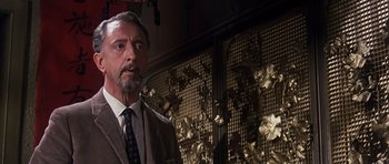 Movie still from “Gambit” (1966), directed by Ronald Neame – A man in a suit and tie standing in front of a wall; Close Up shot, Low angle