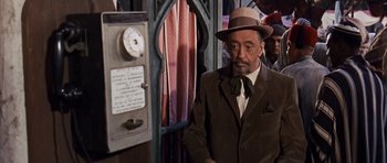 Movie still from “Gambit” (1966), directed by Ronald Neame – An older man wearing a suit and a hat standing in front of an electric meter; Medium shot, Low angle