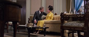 Movie still from “Gambit” (1966), directed by Ronald Neame – A man and a woman sitting on a couch in front of a coffee table; Medium shot, Over the shoulder angle