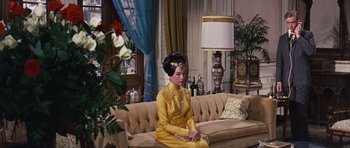Movie still from “Gambit” (1966), directed by Ronald Neame – A woman sitting on top of a couch in a living room; Wide shot, High angle
