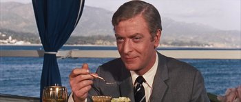 Movie still from “Gambit” (1966), directed by Ronald Neame – A person sitting at a table with a plate of food; Close Up shot, Over the shoulder angle