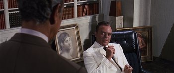 Movie still from “Gambit” (1966), directed by Ronald Neame – A man sitting in front of a painting in a room; Medium shot, Over the shoulder angle