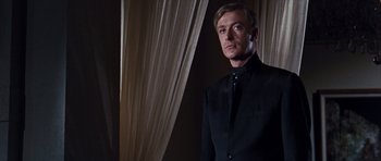 Movie still from “Gambit” (1966), directed by Ronald Neame – A man in a black suit standing in front of a curtain; Medium shot, Low angle