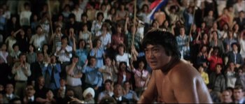 Movie still from “Game of Death” (1978), directed by Robert Clouse – A crowd of people in a stadium with a man in the foreground; Medium shot, Low angle