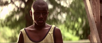 Movie still from “George Washington” (2000), directed by David Gordon Green – A young man in a yellow tank top is standing in front of trees; Close Up shot, Over the shoulder angle