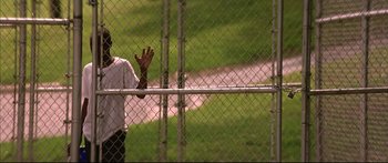 Movie still from “George Washington” (2000), directed by David Gordon Green – A man holding his hand out behind a chain link fence; Medium shot, Low angle
