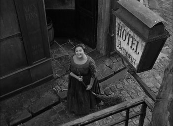 Movie still from “Gervaise” (1956), directed by René Clément – An old photo of a woman holding an umbrella in front of a hotel; Wide shot, High angle