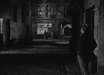 Movie still from “Gervaise” (1956), directed by René Clément – A black and white photo of a man and a woman sitting in a courtyard; Wide shot, High angle
