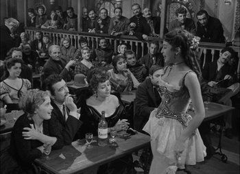 Movie still from “Gervaise” (1956), directed by René Clément – A woman in a corset standing in front of a group of people at a table; Wide shot, Over the shoulder angle