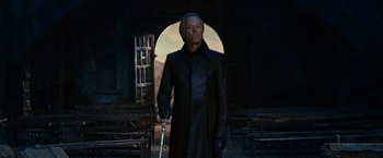 Movie still from “Ghost Rider” (2007), directed by Mark Steven Johnson – A man in a black coat holding a sword in front of an open door; Medium shot, Low angle
