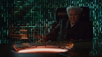 Movie still from “Ghost in the Shell” (2017), directed by Rupert Sanders – An older man sitting at a table in front of a brick wall; Medium shot, Low angle