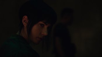 Movie still from “Ghost in the Shell” (2017), directed by Rupert Sanders – A young woman is looking at the camera while a man is standing in the background; Close Up shot, Over the shoulder angle