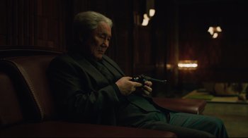 Movie still from “Ghost in the Shell” (2017), directed by Rupert Sanders – An older man sitting on a couch holding a gun; Medium shot, Over the shoulder angle