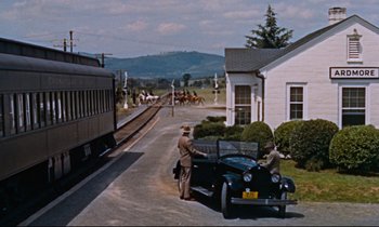 Movie still from “Giant” (1956), directed by George Stevens – An older man standing next to an old car in front of a train station; Extreme Wide shot, Low angle