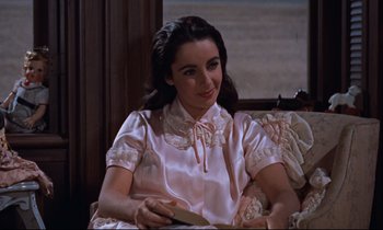 Movie still from “Giant” (1956), directed by George Stevens – A woman in a pink dress sitting on a couch holding a book; Medium shot, High angle