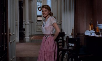 Movie still from “Giant” (1956), directed by George Stevens – A woman in a pink dress standing next to a chair; Medium shot, Over the shoulder angle