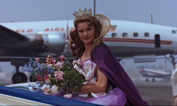 Movie still from “Giant” (1956), directed by George Stevens – A beautiful young lady wearing a tiara and holding a bouquet of flowers in front of an airplane; Medium shot, Low angle