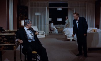 Movie still from “Giant” (1956), directed by George Stevens – A man sitting in a chair in front of two other people; Wide shot, Over the shoulder angle
