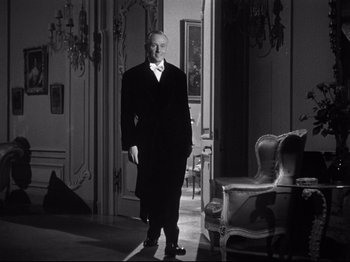 Movie still from “Gilda” (1946), directed by Charles Vidor – A man in a suit and tie standing in front of a chair; Medium shot, Low angle