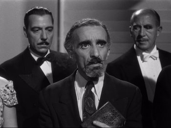 Movie still from “Gilda” (1946), directed by Charles Vidor – A man in a suit and tie holding a book in front of two other men; Medium shot, Low angle