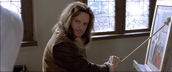 Movie still from “Girl with a Pearl Earring” (2003), directed by Peter Webber – A man sitting in front of a stained glass window holding a pen; Close Up shot, Over the shoulder angle