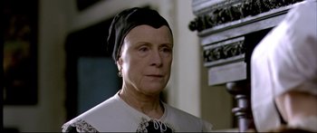Movie still from “Girl with a Pearl Earring” (2003), directed by Peter Webber – An older woman wearing a black hat and a white dress; Close Up shot, Low angle
